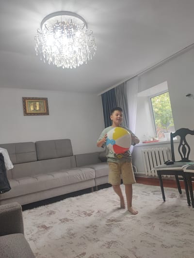 A young boy stands in a living room holding a large, inflatable beach ball. He is wearing a light green t-shirt with a graphic print and tan shorts. The beach ball is multicolored, featuring sections of yellow, red, blue, and white.
The room is well-lit, with natural light streaming in from a window to the right. The window is adorned with sheer white curtains and darker grey drapes. Outside the window, trees and greenery are visible, suggesting it is daytime. A white radiator runs along the wall beneath the window.
In the foreground, a patterned rug with a cream base and abstract brown and white designs covers the floor. A portion of a large, modular grey sofa is visible on the left side of the image, with a smaller grey armchair in the lower left corner. A framed piece of artwork, possibly religious given the text within, hangs on the wall above the sofa.
A prominent crystal chandelier hangs from the ceiling, casting a bright light. To the right of the boy, two dark wooden chairs with upholstered seats are partially visible. The scene appears to be set in a home in Ayagöz, Kazakhstan. The boy's expression suggests he is engaged in play or is about to start.