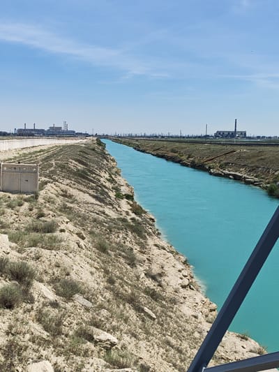 An outdoor, eye-level, long shot captures an industrial canal in the city of Ömirzaq, Kazakhstan, on a clear day under a bright blue sky with scattered clouds. The canal, a prominent turquoise waterway, dominates the right side of the frame, its banks lined with rocky, sparsely vegetated terrain. The left bank features a concrete retaining wall and a large, rectangular concrete structure with a decorative pattern of holes, possibly a ventilation shaft or utility box. Beyond the canal and retaining wall, industrial structures, including smokestacks and buildings, are visible in the distance, indicative of an industrial area. The foreground shows a metal railing, suggesting the viewpoint is from a bridge or elevated platform overlooking the canal. The overall scene conveys a sense of industrial infrastructure within a natural, arid environment.