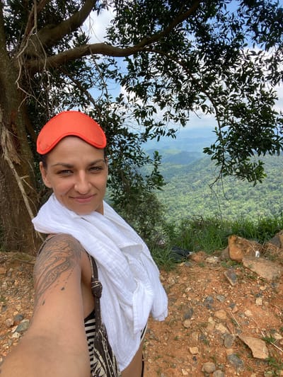 In a selfie taken outdoors with a backdrop of lush green mountains and a cloudy sky, a woman with dark hair pulled back and an orange eye mask on her forehead smiles at the camera. She wears a white scarf draped around her neck and shoulders, and a dark patterned strap of a bag is visible. Her left arm, adorned with a large tattoo, is extended as she holds the camera. The foreground consists of reddish-brown soil with scattered rocks and patches of green grass. The location is identified as the city of Phan Son, Vietnam.