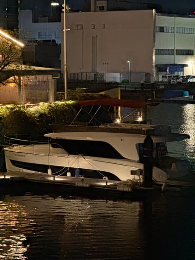 At night, a yacht is docked at a pier on the side of a canal in Shinagawa, Japan. The yacht is white with black trim. Its name, "SUPERIOR II", is visible on the hull. The canal water is dark and reflective, shimmering from city lights. Buildings line the opposite side of the canal, including a multi-story structure with windows and a commercial building with signage. Foliage grows between the canal and the buildings, illuminated by ground-level lighting. A lamp post is visible on the left side.