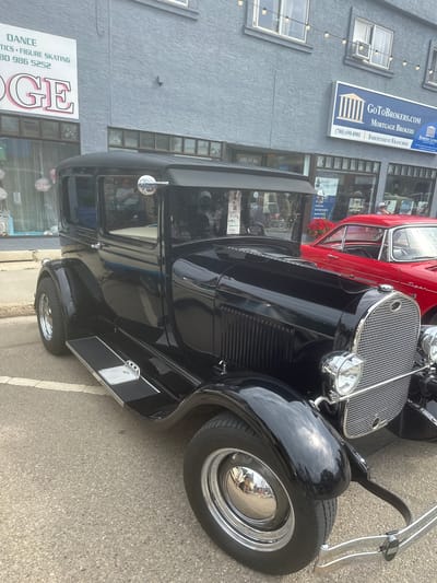 This media file captures a street view featuring a classic black car parked next to a red vintage car. The black car, likely from the 1930s, is the primary focus, showcasing its gleaming chrome accents, prominent grille, and polished black bodywork. The red car is visible behind it, also appearing to be a vintage model.
The background reveals a building with a grey textured facade. Signage on the building indicates commercial businesses, including "GoToBrokers.com Mortgage Brokers" with a phone number and "Dominion Lending Centres." Another sign below announces "DANCE TICS. FIGURE SKATING" with a phone number. The street appears to be paved, with white lane markings visible.
The scene suggests a daytime setting, with natural light illuminating the vehicles and buildings. The overall atmosphere is one of a casual outdoor gathering or event, possibly a car show or a local street event in the city of Leduc, Canada, as hinted by the address "4816-50 Ave." and the business names. No specific activity or event is explicitly depicted, beyond the presence of the cars and businesses. The reflections in the car's chrome and paint suggest a clear sky with some clouds.