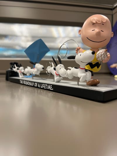 A close-up, eye-level shot shows a figurine display featuring Charlie Brown and Snoopy dogs in a procession. Charlie Brown, bald with a yellow shirt featuring a black zigzag pattern, smiles with closed eyes and holds a small brown nut in his right hand. A metal wire loop extends from his left hand, and a Snoopy dog with black ears and a red collar runs alongside him, looking forward. Behind this Snoopy, a line of three more Snoopy dogs, all with red collars, follows. The first Snoopy in the line is a figurine with its mouth open, as if in mid-stride. The second Snoopy is also in motion, and the third is partially obscured by the others. A small blue object, possibly a folded umbrella, stands behind the first Snoopy. The figurines are positioned on a black base with white text that reads "THE FRIENDSHIP OF A LIFETIME." The display sits on a light brown, reflective surface. In the background, vertical blinds are visible behind a horizontal dark band. The overall scene is a still display, likely indoors, with no discernible city or specific location.