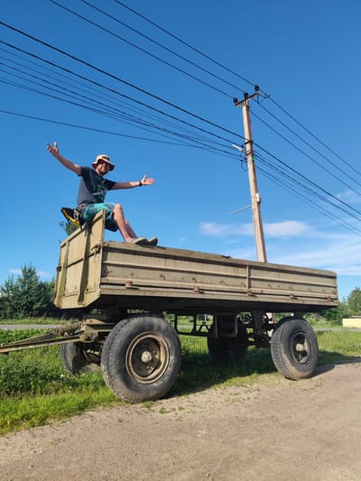 In Zashchita, Russia, a man wearing a bucket hat and shorts sits on the edge of a large, olive-green trailer, his arms outstretched as if in greeting or celebration. The trailer is parked on a dirt path next to a grassy area. Power lines are visible overhead against a clear blue sky, with a wooden utility pole nearby. The man appears to be chewing on something, possibly gum or a piece of candy, which is creating a visible stream of bubbles or strings. The trailer has large, rugged tires and appears to be old or industrial. The background shows greenery and trees, suggesting a rural or suburban setting outside of the main city center.