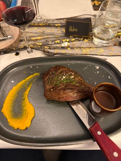 A steak rests on a gray plate with a swipe of orange sauce. It's seasoned with salt and herbs. A small bowl of gravy sits beside it, alongside a steak knife. The table setting includes a wine glass filled with red wine, a water glass, and a name card that says "Pedro". A patterned table runner lies beneath the plate.