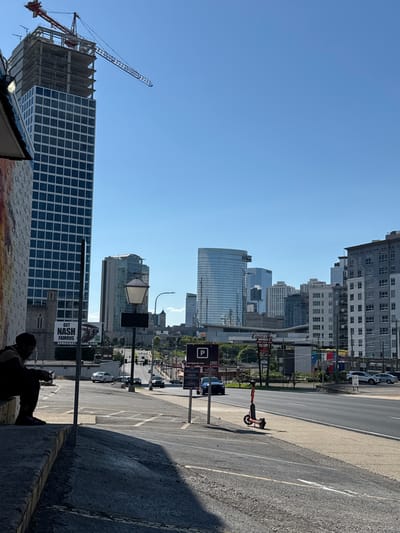 This is a street-level, eye-level photograph captured on a sunny day in Nashville, Tennessee, USA, showing a cityscape with a prominent construction site in the foreground. On the left, a person is seated on concrete steps, looking down, partially obscured by shadow. Adjacent to them is a building with a brick facade adorned with a mural on the left.
Dominating the background is a tall, blue-windowed skyscraper under construction, topped with a red crane. Several other modern buildings, varying in height and architectural style, form the city skyline. A street with several cars driving on it stretches into the distance. Signs for "GET NASH FAMOUS" and "USED CARS" are visible on billboards and building facades. A white street lamp stands near the center of the frame, and a red electric scooter is parked on the sidewalk to the right. The overall impression is one of urban development and city life under a clear blue sky.