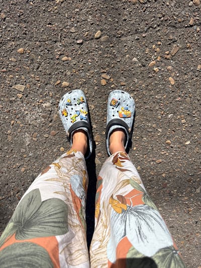 An overhead view captures the lower legs and feet of an individual standing on a dark, granular ground surface in Kropachyovo, Russia. The person wears wide-leg trousers featuring a botanical print with large green leaves and abstract orange and brown shapes against a light background. On their bare feet are light grey Crocs-style clogs with black straps. The left clog is adorned with multiple Jibbitz-style charms, predominantly depicting characters from "The Simpsons," including Bart, Lisa, Homer, and Marge. The right clog displays charms including an "N" block and a depiction of "The Simpsons" family seated on a couch. The ground beneath the individual is dark grey, textured asphalt or packed gravel, embedded with numerous small stones and pebbles. Shadows are visible, suggesting bright, direct overhead lighting.