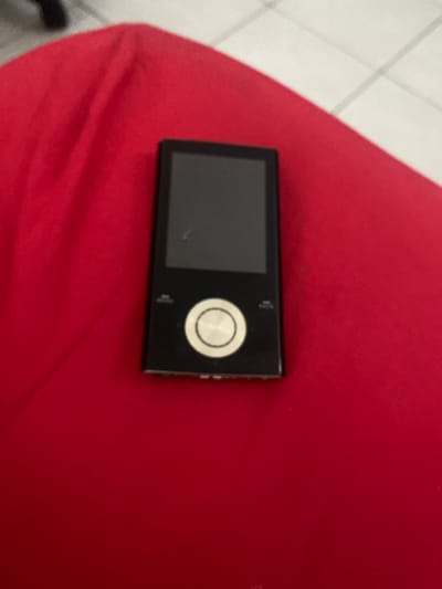 A black digital media player rests on a textured, crumpled red fabric surface in Capesterre-Belle-Eau, France. The device is rectangular, oriented vertically, featuring a dark, blank screen at the top and a circular control dial with a metallic finish below it. Small labels or buttons are visible flanking the control dial. The bottom edge of the device reveals several small apertures, likely ports or grilles. A faint scratch or mark is visible on the screen's surface. In the upper right background, a light-colored tiled floor with visible grout lines is present. No individuals are depicted, and no specific actions or interactions are occurring.