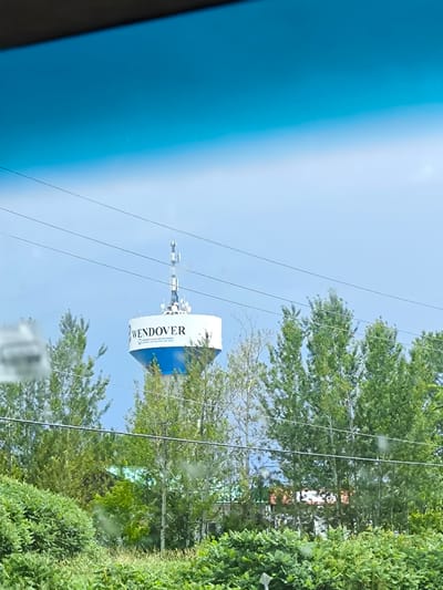 An eye-level, exterior, medium shot captures a water tower with the name "WENDOVER" in black lettering on its white cylindrical upper section. The tower has a blue base and is topped with a communications antenna. It is surrounded by lush green trees and foliage. Power lines stretch across the sky in front of the tower. The scene appears to be in Alfred and Plantagenet, Canada, indicated by the water tower's signage and geographical context. The sky is a clear, bright blue. The image is taken from inside a vehicle, as suggested by the dark tinting at the top of the frame and the slight reflection on the surface. No people or animals are visible in the frame.