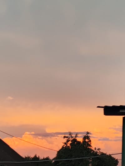 This is a photograph of a sunset over a rural landscape. The sky is filled with soft hues of orange, pink, and gray, with some scattered clouds. In the foreground, there are silhouetted trees and the rooftops of buildings. A power line stretches across the frame. The scene evokes a sense of tranquility and the end of the day. The time of day is clearly sunset. The location cues of buildings and trees suggest a residential or semi-rural setting. The overall mood is peaceful and serene.