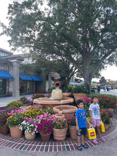 Two children are lying on their sides on a planter decorated with flowers and pottery, holding yellow LEGO bags. The planter is located in front of a building with blue awnings in the city of Lake Buena Vista, United States. A large tree looms over the scene, with other people and structures visible in the background. A fountain is also present, with water flowing from one pot to another. The children appear to be posing for a picture. The location is likely a commercial or recreational area, given the presence of stores, flowers, and other people.