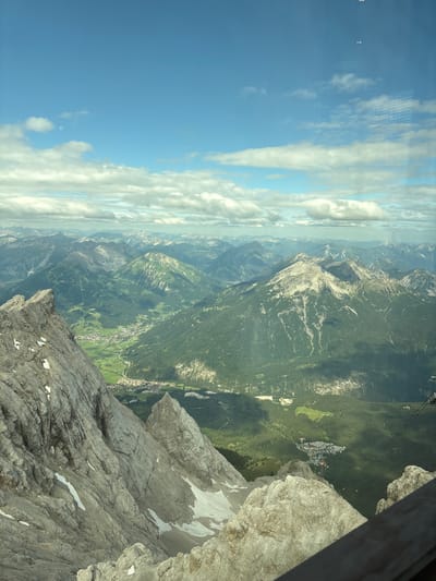 The image captures a panoramic view of the Tyrolean Alps, with a focus on the Zugspitze region near Ehrwald, Austria. In the foreground, rugged, rocky mountain peaks, partially covered with snow, dominate the left side of the frame. These peaks exhibit a rough, weathered texture.

Below these imposing mountains, a verdant valley unfolds, featuring lush green slopes and dense forests. A small town, identifiable by its buildings and infrastructure, is nestled within the valley, with roads winding through it. The landscape recedes into the distance, revealing a series of layered mountain ranges under a sky filled with scattered, fluffy white clouds against a bright blue backdrop.

The perspective suggests the image was taken from a high vantage point, possibly from a cable car or a viewing platform. A glimpse of a wooden railing or structure is visible in the bottom right corner, further supporting this idea. The overall scene is one of natural grandeur, showcasing the dramatic topography of the Alps and the charming human settlement within it.
