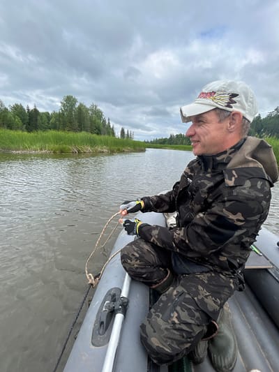 A man in a camouflage jacket and pants, a white baseball cap, and green rubber boots is sitting in a gray inflatable boat on a body of water. He is holding a rope in his hands, and the rope is tied to the boat. The man is looking to his right and smiling. The boat is surrounded by green vegetation and trees on the bank of the water. The sky is overcast. The scene appears to be in a rural or natural setting, possibly near Detskii Dom (Bashenino), Russia, given the natural landscape.
