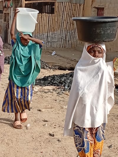 Two individuals are depicted walking on a dirt path in Gayawa, Nigeria, under bright daylight.

The person on the left wears a teal head covering, a blue and gold striped skirt, and light pink sandals. This individual carries a white plastic bucket filled with water on their head, stabilizing it with their right hand, while their left hand shields their eyes. Their face is partially obscured.

The person on the right wears a white head covering that conceals most of their face, exposing only their eyes and forehead, and a patterned skirt with blue, orange, and brown motifs. This individual carries a dark-colored, possibly black, basin or tub on their head. Their gaze is directed forward.

The immediate ground is uneven dirt with scattered small debris. In the middle ground, a pile of dark, charred material is visible. The background features a light-colored structure with a vertical bamboo-slat fence or wall behind the left individual, and a wall with a dark rectangular opening further back on the right.