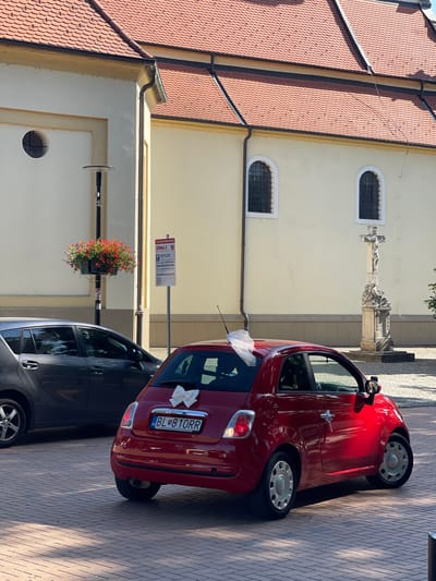 A red Fiat 500 adorned with a white bow on the rear is parked on a brick-paved street in front of a building with a red tiled roof. The car's license plate reads "BL 810RR". A grey hatchback is parked to the left of the Fiat. In the background, the building features cream-colored walls and arched windows. A stone crucifix stands to the right of the building. A hanging planter with red flowers is visible on a pole in front of the building, along with a parking zone sign. The scene appears to be located in Stupava, Slovakia.