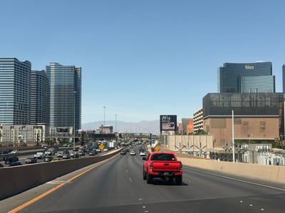 This image depicts a multi-lane highway in Paradise, Nevada, United States of America, with traffic moving in both directions. In the foreground, a red pickup truck is driving in the rightmost lane. The backdrop features several high-rise buildings, including the Vdara Hotel & Spa and buildings that constitute CityCenter, set against a clear blue sky. The Aria Resort & Casino is visible behind a billboard for the same establishment. Mountains are present in the far distance. A concrete barrier separates the highway lanes.