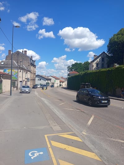 The image depicts a street scene in Nancy, France, under a bright blue sky with scattered white clouds. In the foreground, a blue square with a white wheelchair symbol indicates a disabled parking spot. Yellow diagonal lines are painted on the asphalt, forming part of a road marking. A dark-colored hatchback car, bearing the license plate "CG-782-TJ", is driving on the right side of the road. The street is lined with buildings on the left, including a multi-story building with a poster on its facade. On the right side of the road, a dense green hedge forms a wall. Further down the street, several other cars are parked or driving. A street light is visible on a pole on the left side of the image. The overall environment suggests a sunny day in a residential or commercial area.