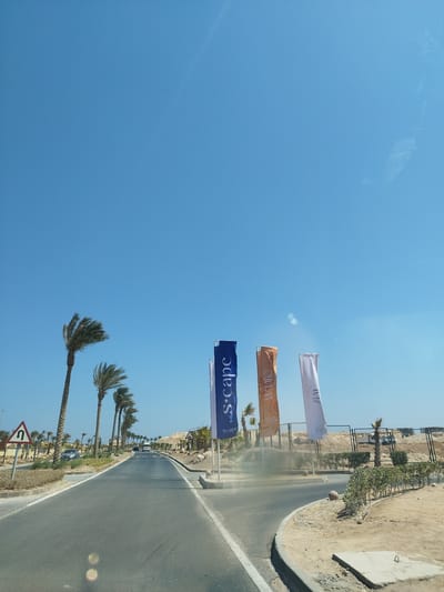 The image depicts a road scene in Ras Abu Soma, Egypt, under a clear blue sky. The road, constructed of grey asphalt, leads into the distance with palm trees lining its sides. On the right of the road, there are three flags displayed: one blue, one orange, and one white. The flags bear the names "S.scape", "WVIE", and "ZUNI" respectively. Behind the flags, there is a fence, some vegetation, and what appears to be construction material or sand. A turning sign is visible on the left side of the road. Cars are seen driving on the road. The image appears to have been taken from inside a vehicle.