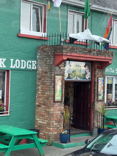 The image shows the exterior of a building in Falcarragh, Ireland, identified by a sign that reads "K LODGE." The building has a bright green textured facade and is adorned with red trim around windows and the entrance. The entrance itself is recessed and framed by a brick facade. Above the entrance, a large window features a colorful, stylized sign that appears to be in Irish script, reading "Coistín na Seamróige." To the left of the entrance, a large framed artwork depicts a woman with flowing red hair. To the right, two smaller framed artworks are visible, one on the brick facade and another on the green wall next to a window.
Flags are visible on the building: a white flag and a green flag are mounted on poles near the roofline, and a rainbow flag is also displayed. In the foreground, a bright green picnic table is set up on the pavement. A dark-colored car is partially visible in the lower right corner of the frame. The environment appears to be an outdoor setting, possibly a street or public area in front of the building. There are potted plants near the entrance and a trash can. The overall impression is that of a pub or guesthouse.