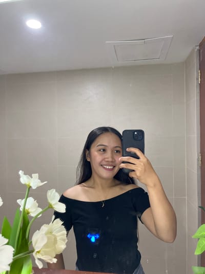 The image is a mirror selfie taken in what appears to be a bathroom. A young woman with long dark hair is smiling at the camera, holding a black smartphone in her right hand. She is wearing an off-the-shoulder black top with a delicate gold necklace featuring a crescent moon pendant. In the foreground, to the left, a bouquet of white flowers with green stems is visible. The background consists of beige tiled walls and a white ceiling with a circular light fixture and an open ceiling access panel. The reflection also shows a brown door frame on the right side of the image, and a portion of a green leafy plant. The overall setting suggests a domestic environment. Based on the user's prompt, the location is identified as Koronadal City, Philippines.