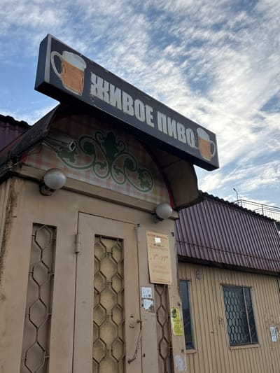 This image depicts the facade of a building, likely a store or pub, located in Ulyanovka, Russia. A prominent sign that says "ЖИВОЕ ПИВО" (Live Beer) in Russian is mounted above the entrance. The sign also features images of beer mugs. Below the sign, the facade is decorated with a checkered pattern and a green floral design. A pair of round lights flank the entrance. A sign with hours and other store information is visible to the right of the entrance. The facade appears slightly weathered. The upper part of the building has windows protected by honeycomb patterned bars. Above the upper facade is a section of corrugated roofing. A blue sky with scattered clouds is visible in the background.