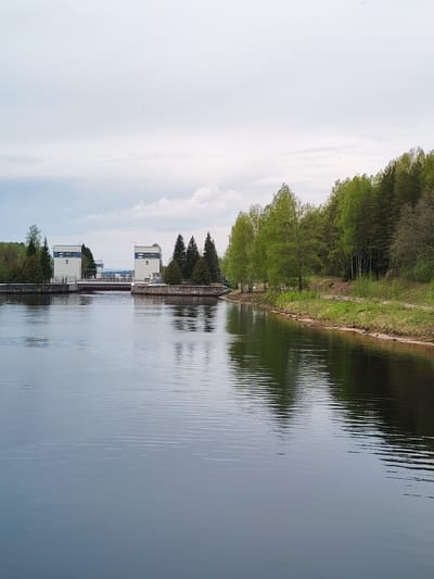 This image shows the Vyacheslavsky Lock ( шлюз Вячеславского) on the Kama River near Alekseevskoe, Russia. The scene features calm water reflecting the sky and shoreline vegetation. Two identical, white, concrete lock towers with blue trim and the numeral "5" are positioned on either side of the lock channel. A forested shoreline with a mix of deciduous and coniferous trees is visible on the right. The sky is overcast.
