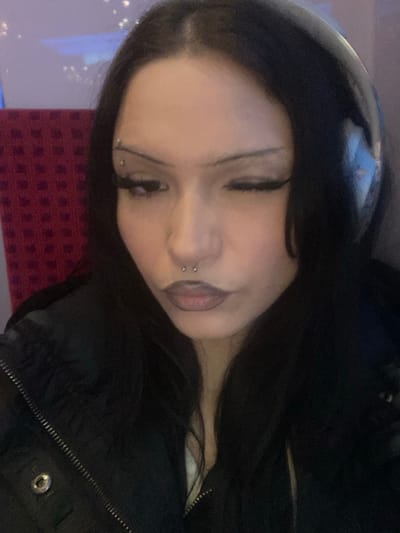 The scene takes place within a public transport vehicle, such as a train or bus, potentially located in or traveling through Enghien-les-Bains, France. The image presents a medium-close-up of a female individual, positioned slightly off-center to the viewer's right. She has long, dark, straight hair, center-parted, and wears light-colored over-ear headphones. Her right eye (viewer's left) is closed in a distinct wink, while her left eye (viewer's right) is open and gazing downwards. Her eyebrows are thin and sharply arched, with a multi-sphered curved bar piercing above her left eyebrow. Dark, winged eyeliner is applied to both eyes, complemented by long eyelashes. A silver-toned horseshoe barbell piercing is visible in her septum. Her lips are pursed, covered with a dark reddish-brown lipstick. She wears a black padded jacket with a visible zipper closure, and a white garment beneath it. The background is blurred, showing dark red, textured fabric consistent with seating upholstery on the left, and lighter-toned surfaces with indistinct bright points of light above it, indicative of an interior cabin environment.