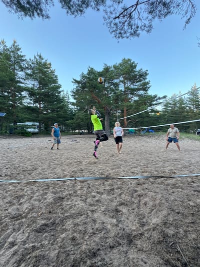 Four individuals are playing volleyball on a sandy court surrounded by pine trees. The court has a net stretched across it. One person in a neon green shirt is jumping to hit the ball. Another person in a blue tank top and shorts stands watching. A woman in a white tank top and black shorts and a man in a tan polo shirt and blue shorts are also on the court. In the background, there are tents and campers, suggesting this might be a campsite or recreational area. The sky is clear and blue. The scene appears to be in a natural outdoor setting with a focus on recreational activity.