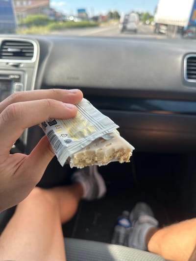 In the image, a hand holds a partially unwrapped snack bar inside a vehicle. The snack bar is light brown with white frosting and appears to be made of granola or oats. The wrapper is silver and white with some text printed on it. The visible text on the wrapper includes "65 g". The scene appears to be set during the daytime, with sunlight coming through the vehicle's windows, casting shadows. The interior of the car is visible, including a dashboard vent and the passenger seat. In the background, through the car window, a blurry outdoor scene is visible, suggesting the vehicle is in motion or parked in an area with buildings and vegetation. A person's legs and feet, wearing sneakers, are visible in the passenger seat. The image conveys a casual moment of someone snacking while traveling. The location is Alès, France.