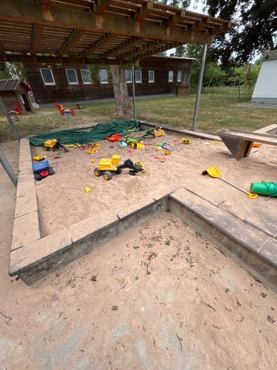 A high-angle, wide shot captures a sandbox filled with various children's toys. The sandbox has concrete borders and is situated in an outdoor setting. The scene is dominated by sand, with a collection of brightly colored plastic toys scattered across it, including trucks, shovels, and a bucket. A large green tarp is partially covering some of the toys on the left side of the frame.
In the background, a wooden building with several windows is visible, surrounded by a grassy area and trees. A wooden, lattice-like structure, possibly part of a playground or a shaded seating area, is prominent on the left side of the image. The overall impression is that of a play area, likely a backyard or park, filled with toys ready for use.
The lighting suggests it is daytime, with no strong shadows indicating a specific time. The weather appears to be overcast or with diffused sunlight, as the light is even and not harsh. There are no people visible in the frame, implying the area is currently unoccupied or the photo was taken at a time when children were not present. The location context of Giengen an der Brenz, Germany, suggests this is a German setting, though no specific landmarks or text confirm this within the image itself.