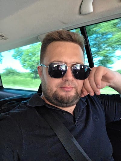 A man wearing sunglasses and a dark blue polo shirt is sitting in the back seat of a car. The man has short brown hair and a beard. He is resting his head on his hand. The window of the car shows a blurry green landscape and blue sky, indicating the car is in motion. A seatbelt is visible across the man's chest. The interior of the car is dimly lit. The scene is captured from a slightly low angle.