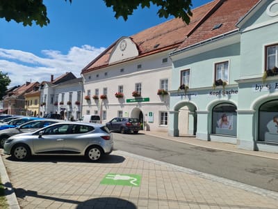 The image depicts a street scene in Sankt Marein bei Neumarkt, Austria, on a sunny day. In the foreground, a silver Renault Clio is parked on a paved area marked with a green electric vehicle charging symbol. To the left of the car, a sidewalk with grass and trees is visible.

The background features a row of buildings with red tiled roofs. The closest building on the right is light blue and has arched entrances with large windows displaying advertisements. Text on these windows includes "Kopf Sache" and "by E". Adjacent to this is a cream-colored building with multiple windows and flower boxes filled with red flowers. A clock face is visible high on this building's facade. Further down the street, other buildings with similar architectural styles are present. Several cars are parked along the street, including a dark-colored SUV. The sky is clear blue with scattered white clouds. The overall scene conveys a tranquil, European town atmosphere.