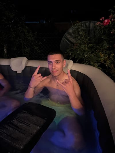 A young man with short hair is sitting in an inflatable hot tub at night. He is wearing a necklace with a cross and making a rock hand gesture with both hands. The hot tub is filled with water that is illuminated by blue and green lights. In the background, there is a dark yard with some greenery and a fence. A rose bush with red flowers is visible on the right side of the image.
