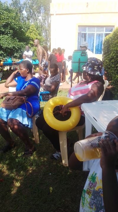 The image depicts an outdoor social gathering in Siaya Township ward, Kenya. In the foreground, an adult female with dark complexion, wearing a blue short-sleeved top and skirt, is seated on a white plastic chair. A brown shoulder bag rests on her lap. To her right, another adult female, also with dark complexion, wears a pink and white horizontally striped sleeveless top and a multi-colored bucket hat, holding a yellow inflatable swim ring on her lap while seated on a white plastic chair beside a white plastic table. A child, partially visible in the bottom right, holds a transparent plastic cup containing an orange liquid.
In the midground, an adult male with dark complexion stands shirtless, wearing patterned swim trunks. Another adult male in a white t-shirt is visible nearby. Several other individuals are present in the background, standing and conversing. A large green plastic barrel, possibly a water dispenser, is positioned on a stand near a light-colored building with blue-framed windows. The environment consists of a sunny day with a clear sky, a grassy lawn, and numerous green trees and bushes providing shade in the background and along the left edge of the frame. The overall scene suggests a casual recreational gathering.