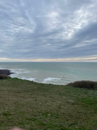The image depicts a coastal scene in Audinghen, France, under an overcast sky. The horizon line is visible across the mid-section, separating a grey, heavily clouded sky from the sea. The sea exhibits a greenish-grey hue with visible whitecaps and waves breaking, indicating active water movement. To the left, a dark, rocky shoreline is present where waves are impacting the land. The immediate foreground consists of a gently sloping, irregularly textured expanse of grass, varying in shades of green and brown, consistent with a cliff edge or elevated coastline. Dense, dark green to brown shrubbery is visible along the top edge of this grassy area, particularly prominent on the right side and also near the rocky shoreline on the left. No individuals, animals, or man-made structures are discernible within the primary landscape. A small, blurred, light-colored object, possibly a finger, slightly encroaches into the lower-center edge of the frame, obscuring a minimal portion of the grass.