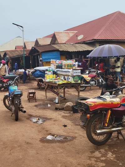 The image depicts an outdoor market scene in Tangu, Nigeria, on an overcast day. Several stalls are set up, selling various goods, predominantly what appear to be agricultural chemicals or similar bottled products. Motorcycles are parked throughout the market, some serving as makeshift display stands. A few individuals are visible, some attending to the stalls, others conversing, and one person is seated on a motorcycle in the foreground. The ground is dusty and uneven, with some puddles of water indicating recent rain or a damp environment. The architecture consists of open-fronted structures with corrugated metal roofs, typical of informal market settings. The overall atmosphere suggests a busy, functional marketplace.