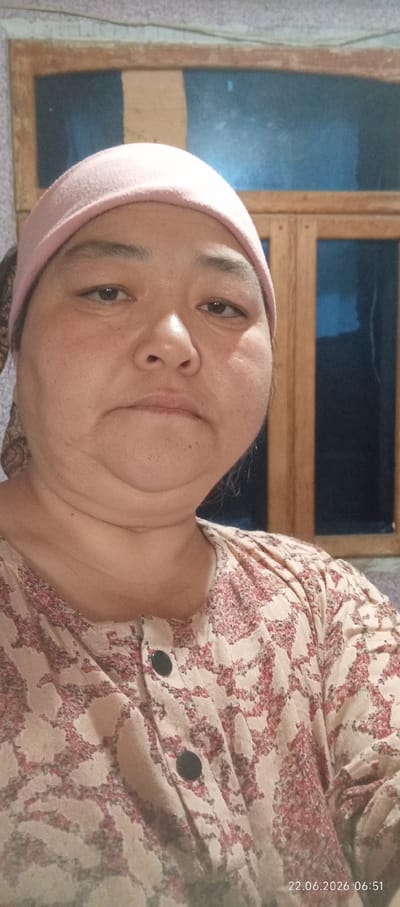 A woman wearing a pink headscarf and a patterned shirt is shown in a close-up, front-facing portrait. She has a neutral expression and appears to be indoors, with a window and a wall visible in the background. The image is dated 22.06.2026 at 06:51.