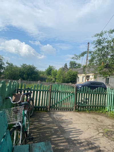 This image depicts a static outdoor scene in Ihnatpil, Ukraine, characterized by a semi-rural or village environment under a partly cloudy sky.
In the foreground, a weathered teal-green wooden bench or low structure is partially visible on the bottom left. Adjacent to it, two bicycles are parked upright next to a green wooden picket fence. The closer bicycle features a green frame, silver fenders, chrome handlebars, and a large, square silver wire basket mounted on the front. The further bicycle, with a red frame, has a brown wicker basket attached to its handlebars. Both bicycles cast long shadows extending towards the lower left.
A prominent green wooden picket fence with a matching gate spans horizontally across the midground. The gate appears slightly ajar. The ground in front of the fence is a light grey paved surface, cracked in various places, with small patches of grass growing through. Long, distinct shadows from the fence pickets fall across this paved area, indicating sunlight from the right side of the frame. A round, dark metal manhole cover is visible embedded in the pavement in the lower right corner.
Behind the green fence, an older, more weathered wooden fence with concrete posts delineates another boundary. Beyond this, dense green foliage and numerous trees form a natural backdrop, suggesting overgrown areas or a small forest.
In the background on the right, a light-colored, single-story house with a grey pitched roof is partially visible. The house has white-framed windows and decorative white trim. A dark blue SUV is parked in front of the house, largely obscured by the green fence. To the right of the house, a wooden utility pole with multiple overhead wires extends upwards. Branches with green leaves from an unseen tree overhang the upper right portion of the scene.
The upper portion of the image reveals a light blue sky with scattered white cumulus clouds. Several thin, dark lines, likely power or communication cables, traverse the sky horizontally. A few tiny, dark specks, possibly birds, are visible in the distant sky. No people are present in the scene, and no active interactions or movements are depicted.