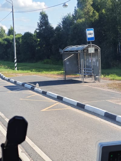 The image depicts a roadside scene in Kuzhlevo, Russia, featuring a bus stop. A bus stop shelter with a bench and a trash can is visible, along with a blue bus stop sign with a white bus icon. The area is paved, with a road marked by yellow lines and a black and white striped curb. A plastic bottle is lying on its side on the curb. In the background, there are trees and a cloudy sky. A utility pole with a lamp is also present. The lower part of the image shows a black object, possibly a camera or part of a vehicle, in the foreground.