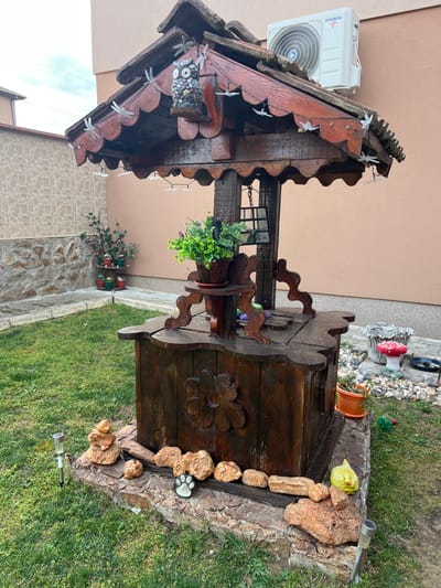 This image depicts an ornamental wooden wishing well situated in a residential garden in Mitino, Bulgaria. The well, constructed from dark-stained wood, features a six-sided shingled roof, supported by carved posts. A string of clear dragonfly-shaped lights is strung along the underside of the roof eaves. A decorative owl figure, composed of multicolored stones, is affixed to one of the roof supports. Below the roof, a dark-colored lantern hangs from a central wooden mechanism. An artificial green plant in a brown pot rests on a small wooden shelf attached to one of the well's supports.
The well's base is cylindrical with a carved floral pattern, resting on a lower stone-faced concrete foundation. This foundation is adorned with various natural-colored rocks, a black and white paw print stepping stone, and two metallic solar garden lights. The immediate surroundings consist of a well-maintained green lawn.
In the background, to the left, a multi-tiered stand holds several potted plants against a light-colored, possibly tiled, wall and a lower stone-faced wall. To the right of the well, a gravel patch contains a red and white mushroom-shaped ornament, a yellow gnome-like figure, and a small orange potted plant. Further back, a light peach-colored building wall is visible, with a white 'KAISAI' brand air conditioning unit mounted high on it. The sky above is overcast. No individuals are present in the scene.