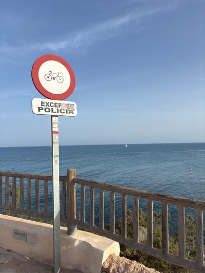The image displays a street sign with a circular red and white sign prohibiting bicycles, followed by a rectangular sign reading "EXCEPTO POLICIA" which translates to "EXCEPT POLICE." The signs are mounted on a metal pole. The background features a calm blue ocean under a bright blue sky with sparse white clouds. A small white sailboat is visible on the horizon. A wooden railing runs along the foreground, bordering a stone wall and some low-lying green bushes. The location is identified as Urbanización Cabo Roig, Spain. The scene is static and no people are present.