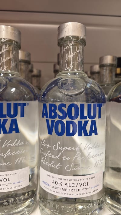 A close-up, eye-level shot shows several bottles of Absolut Vodka arranged on shelves. The bottles are made of clear glass and are filled with a clear liquid. The labels are white with blue lettering. The closest bottle, on the right, has "ABSOLUT VODKA" printed in large blue letters. Below that, in white script, it reads "This Superb Vodka is Crafted to Perfection, Absolut Since 1879." A smaller white label on the bottle states "MADE WITH SWEDISH WATER & WINTER WHEAT 40% ALC/VOL IMPORTED PRODUCED & BOTTLED IN THE VILLAGE OF ÅHUS, SWEDEN." The bottle also features an embossed portrait of a man in a circular medallion. The background is blurred, but appears to be shelving in a store. There is no indication of the city of Līvāni, Latvia in the image.