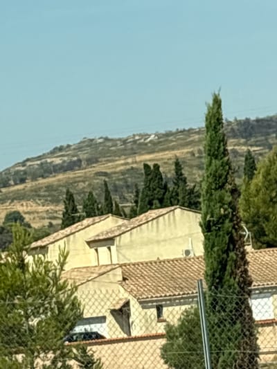 The image depicts a rural or suburban scene with buildings and natural elements. In the foreground, a chain-link fence is visible, with green foliage on the left. Behind the fence, parts of buildings with terracotta tiled roofs and light-colored walls are apparent. An air conditioning unit is mounted on one of the walls. Several tall, slender cypress trees are interspersed with other green trees, creating a vertical element against the landscape. In the background, a sloping, dry, and barren hillside is visible under a clear blue sky. The overall impression is one of a sun-drenched environment, likely in a Mediterranean climate. The location is identified as Bages, France, suggesting a provincial or countryside setting within that region.