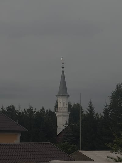The image shows a white minaret with a crescent moon and star on top, situated behind a dark, dense line of evergreen trees. In the foreground, the roofs of several buildings are visible, one with a dark, tiled surface and another with a yellow wall section. The sky is overcast and gray, suggesting a cloudy day. The overall scene appears to be a residential or semi-rural area. Based on the context, this minaret is located in the city of Nurkeevo, Russia.