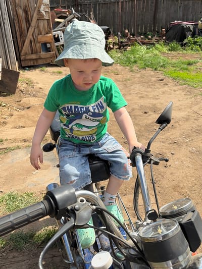 The image shows a young child, likely a toddler, sitting on a motorcycle outdoors. The child is wearing a green t-shirt with a shark design, ripped denim shorts, a light-colored hat, and patterned socks. The motorcycle is a smaller model, with a black seat, a chrome handlebar, and gauges on the front. The background shows a dirt ground, wooden fences, and some vegetation. Considering the presence of rural fencing and the lack of modern structures, the location in Ul'yanovka, Russia, is most likely a rural area on the outskirts of the city.