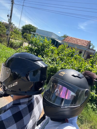 Two individuals wearing helmets are seated on a vehicle, facing forward, with a rural landscape in the background. The person on the left wears a black LS2 helmet and a grey and black checkered shirt. The person on the right wears a black helmet with the brand name "Royal" in gold script and a pink-tinted visor. They are positioned in front of dense green foliage and a light blue building with a red-tiled roof. Power lines are visible against a clear blue sky, and a utility pole stands to the left. The location is identified as Hong Son, Vietnam.