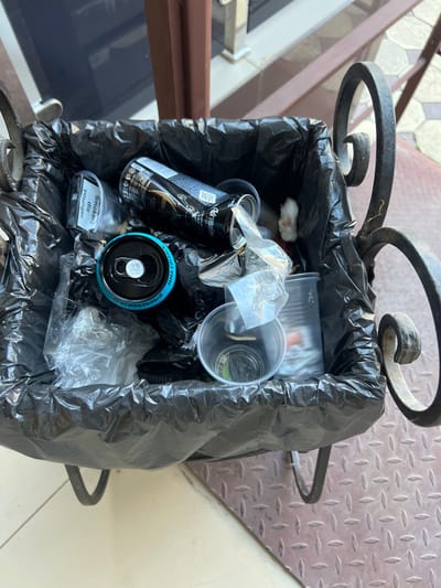 An overhead, medium shot captures a black trash bag overflowing with discarded items inside a decorative metal trash bin. The bin is situated outdoors on a textured, diamond-plate metal surface. Among the visible trash are a black energy drink can with gold accents, a blue-lidded can with Cyrillic text that reads "ОРИГИНАЛЬНЫЙ" (Original), and several clear plastic cups. A small white object, possibly cotton, is also visible within the trash. In the background, a dark blue surface, likely a door or window, and a brown railing are present, with a glimpse of a tiled walkway. The scene appears to be in Magas, Russia, based on the Cyrillic text.