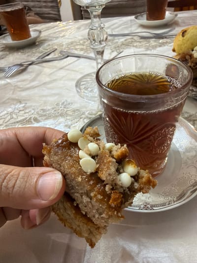 A close-up shot captures a hand holding a piece of a sweet pastry, possibly a cookie or bar, topped with white chocolate chips and a caramel glaze. In the background, a glass of dark tea sits on a decorative saucer, and a portion of a crystal wine glass is visible. The table is covered with a white tablecloth, and cutlery can be seen on the left side of the frame. The scene appears to be set in Balakənd, Azerbaijan, suggesting a domestic or cafe setting where tea and pastries are being enjoyed.