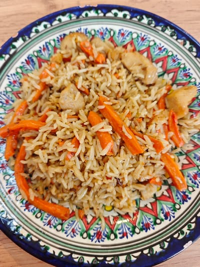 A close-up overhead shot shows a colorful, patterned plate filled with pilaf. The pilaf is composed of long-grain rice, julienned carrots, and pieces of light-colored meat, possibly chicken. The plate has a dark blue rim with an intricate design of green, red, and white geometric patterns and stylized floral motifs. The pilaf is piled high in the center of the plate, with the carrots and meat interspersed throughout the rice. The surface underneath the plate appears to be a light-colored wooden tabletop. The image is sharply focused on the food and the plate, with a shallow depth of field that blurs the background slightly. There are no people visible in the image, and no identifiable landmarks or specific details that would indicate a location in Cheremushki, Russia. The scene is a still life of food.