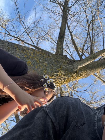 The image, likely taken in Beli Brod, Bulgaria, shows a low-angle shot of a scene near a large tree. A young woman is centrally positioned, wearing a flower crown of daisies in her long brown hair. She wears a black t-shirt and a silver bracelet, partially covering her face with her hands. To the right is a person in a dark denim jacket, obscuring part of the view. The large tree is the primary backdrop, with mossy bark and bare branches reaching towards a blue sky. The image suggests a casual, outdoor setting, possibly a park or natural area.