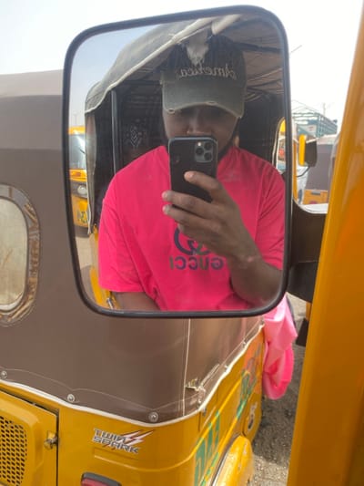 This image depicts a male individual taking a selfie in the rearview mirror of a yellow tricycle in Rijiyar Lemo, Nigeria. The individual, seen from the chest up, is wearing a distressed olive-green baseball cap, partially obscuring his face. Light-colored hair or material protrudes from a hole in the top of the cap. He wears a bright pink short-sleeved t-shirt with a prominent blue and green "GUCCI" logo displayed reversed on the front. His right hand holds a dark-colored smartphone vertically, with the triple camera module visible, actively capturing the reflection. A light-colored scar is visible on his right forearm. The mirror itself is attached to a yellow three-wheeled vehicle (a Keke Napep or tuk-tuk) featuring a brown fabric canopy secured by rivets. The vehicle's side panel displays "TWIN SPARK" branding with a lightning bolt graphic. In the reflected background, other yellow vehicles are visible under bright daylight conditions, suggesting an outdoor urban or market setting.